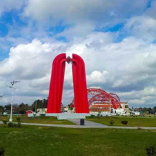 Two tall red arches forming a doorway-like sculpture in a grassy park under a blue sky with clouds, connected at the top by a decorative element.