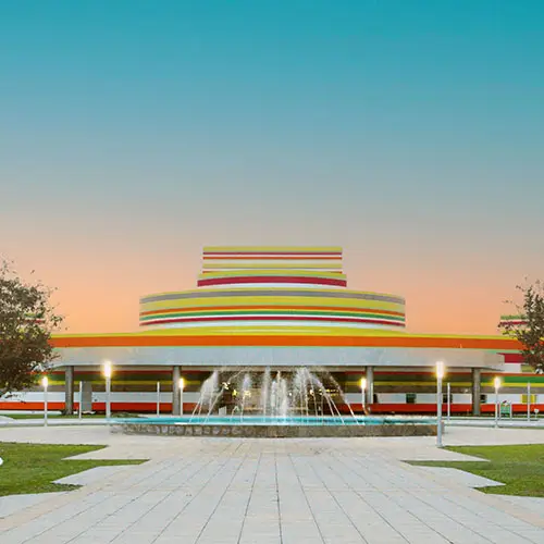 Rainbow-striped circular building with stacked rings and a water fountain in front at sunset.